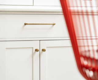 A close-up of white cabinetry featuring brass handles, with a hint of a red wire chair in the foreground.
