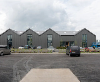 Modern gray building with a distinct peaked roof, surrounded by parked cars and greenery under a cloudy sky.
