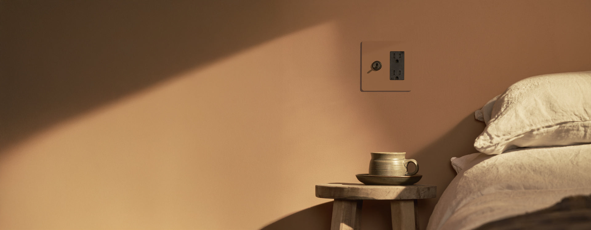 A cozy bedside scene featuring a simple wooden stool with a ceramic cup and saucer, next to a wall-mounted power outlet.
