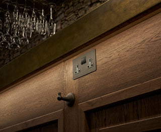 Close-up of a wooden bar counter featuring a power outlet and a hook, with a backdrop of hanging glasses.
