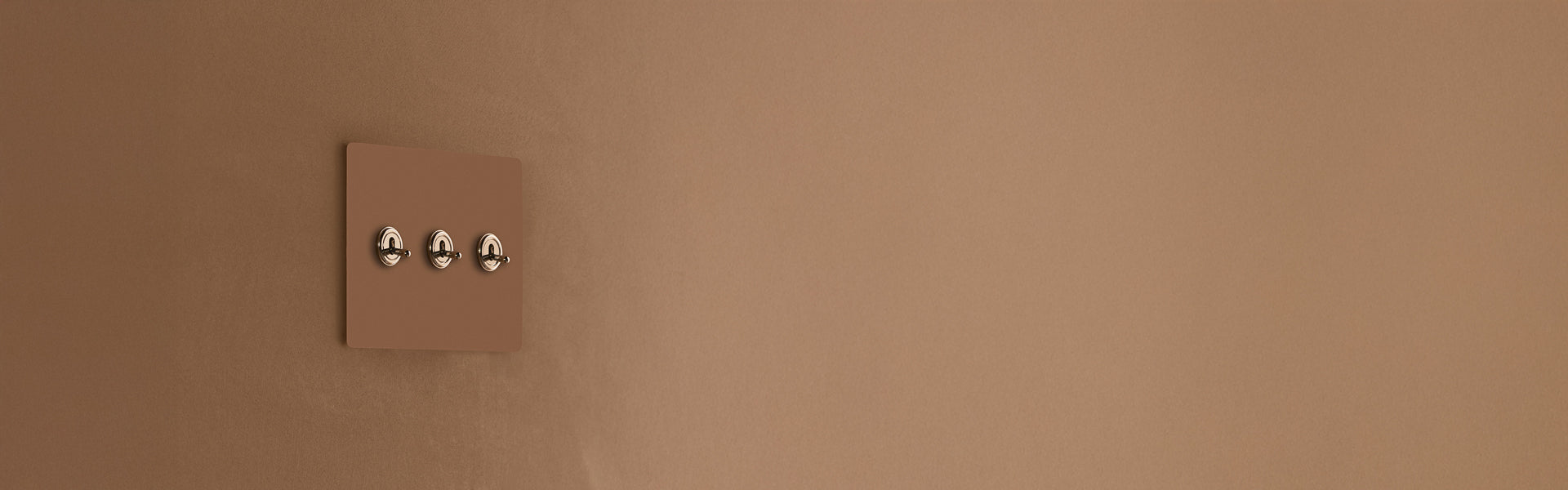 A modern light switch panel with three metallic switches on a smooth, warm-toned brown wall.
